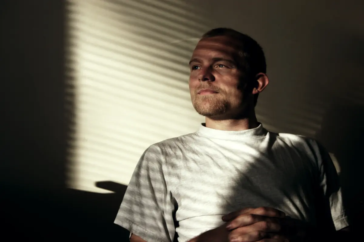 A man sat looking out a window with shadows on him.